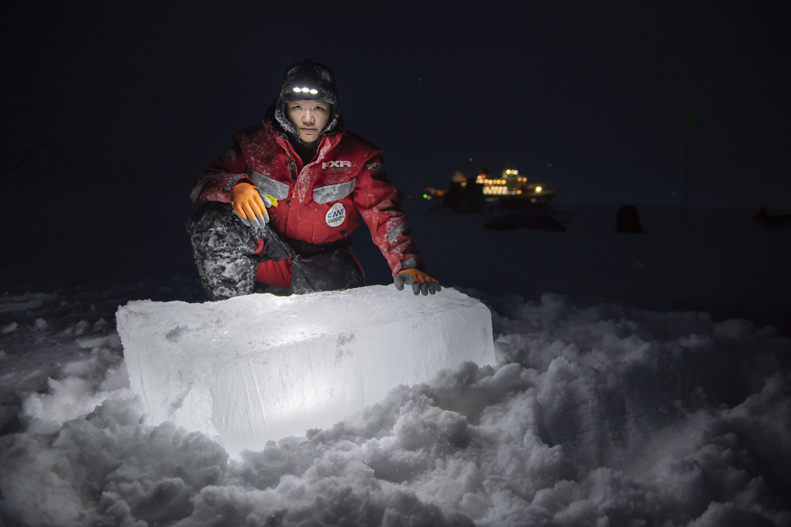 Forscherin Allison Fond vor einem Eisklotz in der Arktis bei Nacht. Das Forschungsschiff ist im Hintergrund zu sehen. Foto: Esther Horvath
